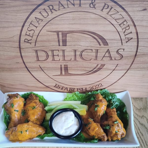 Buffalo wings with ranch dressing and celery sticks on a plate, with the Delicias Pizzeria logo in the background.