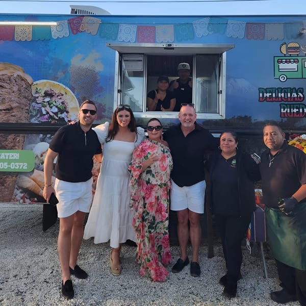 Six people, including restaurant staff, pose in front of the Delicias Sobre Ruedas food truck.