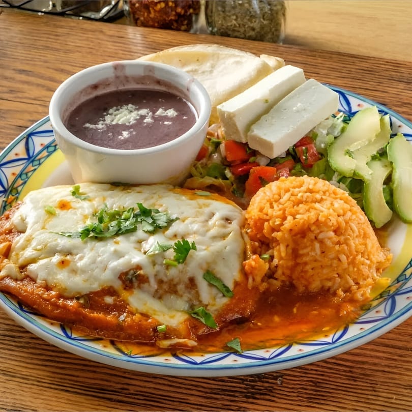 A plate of enchiladas with melted cheese, rice, black beans, avocado, and fresh cheese.