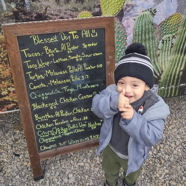 A young child in a winter hat gives a thumbs-up next to a Delicias Sobre Ruedas menu board.