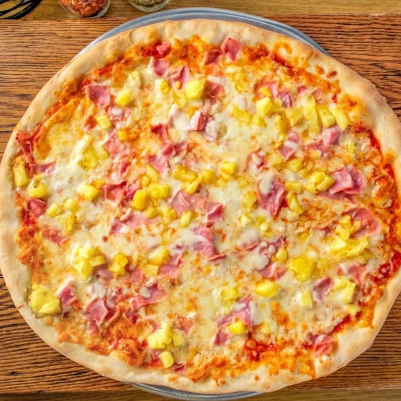 A Hawaiian pizza from Delicias Sobre Ruedas with ham, pineapple, and melted cheese on a wooden table.